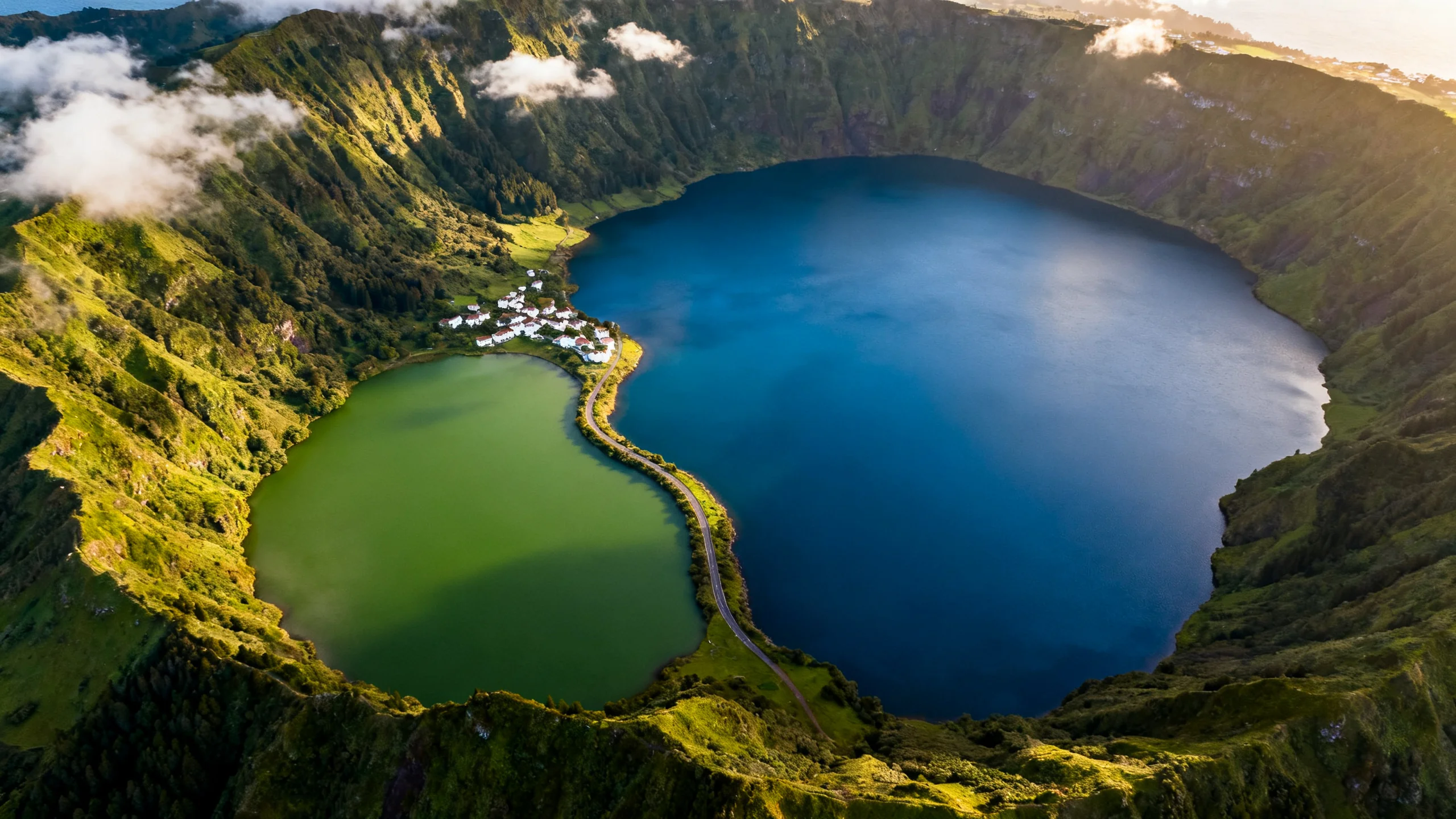 Aerial view of the twin crater lakes of Sete Cidades on São Miguel, the green and blue lakes joined by a narrow strip of land, with steep volcanic crater walls rising on all sides and a few scattered clouds in the sky