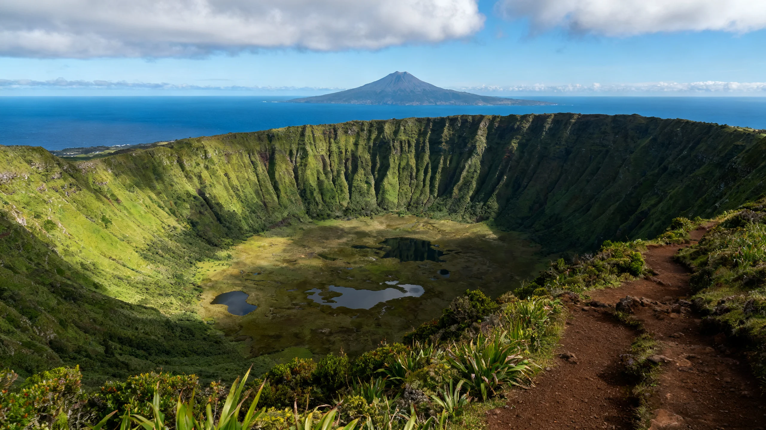 The Caldeira do Faial seen from the rim trail, a 2 kilometre wide volcanic crater at 1,000 metres altitude with steep green-vegetated walls dropping to a wetland floor, the deep blue Atlantic ocean and the Pico volcano visible in the distance on the horizon under a partly cloudy sky