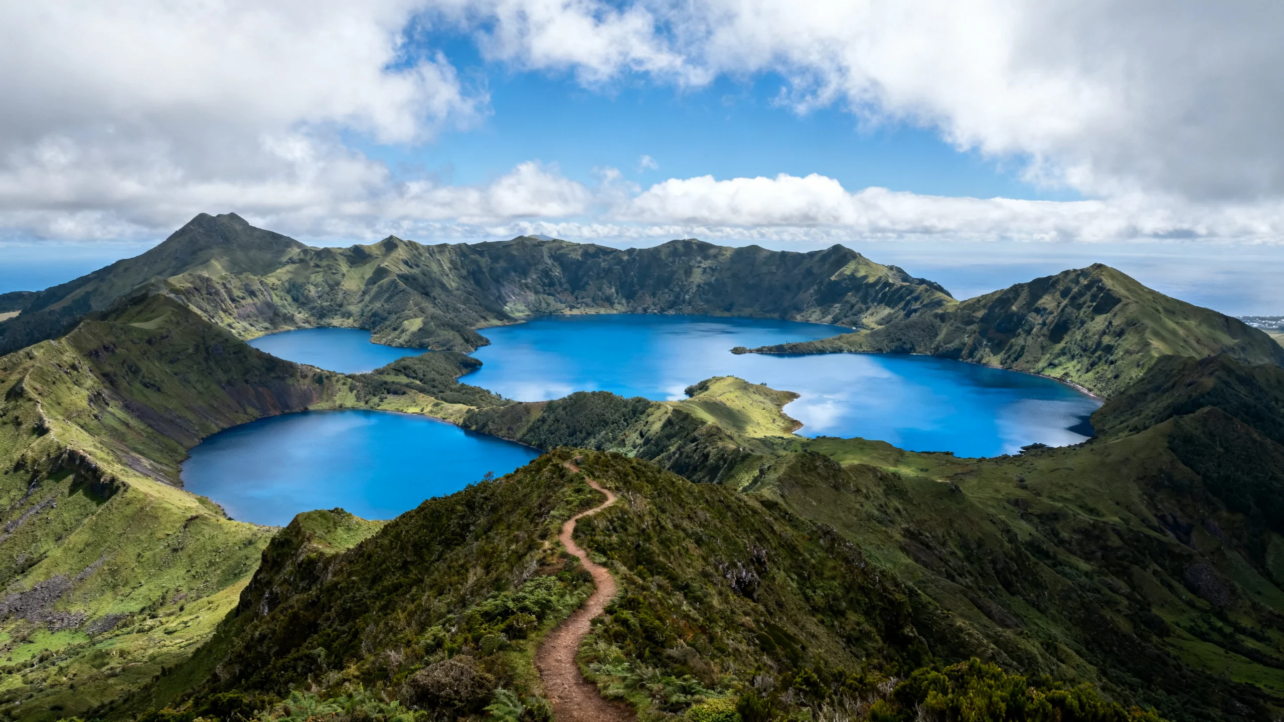 The seven crater lakes of the central Flores plateau seen from the Miradouro do Pico Alto viewpoint, with several lakes visible as bright blue mirrors set in the green volcanic landscape under a partly cloudy sky, the surrounding peaks rising gently around the lake basins